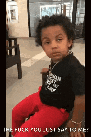 a little boy wearing a black shirt and red pants is sitting on a chair and making a funny face .