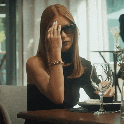 a woman wearing sunglasses sits at a table holding a glass of wine