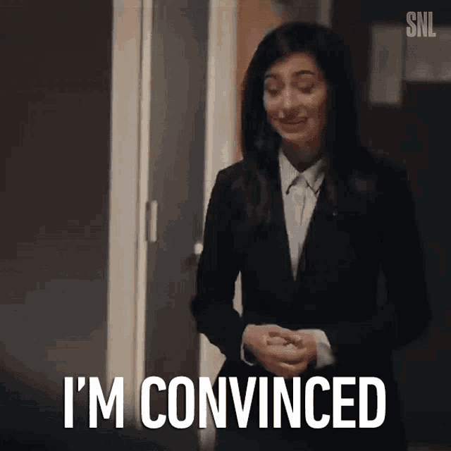 a woman in a suit is standing in front of a door and says i 'm convinced