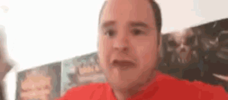 a man in a red shirt is making a funny face while sitting in front of a wall .