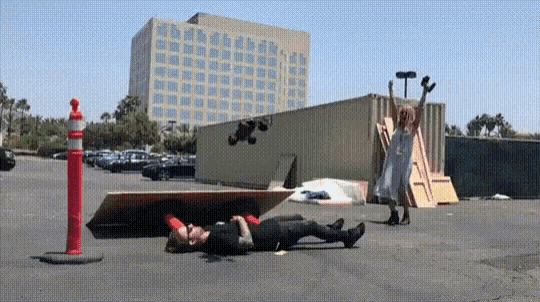 a man is laying on the ground in a parking lot while a woman stands behind him holding a hammer