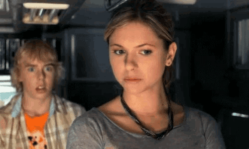 a woman wearing a necklace looks at the camera with a man behind her