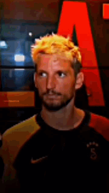 a man with yellow hair and a beard is standing in front of a red letter a