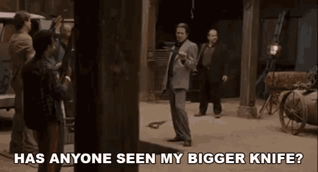 a man in a suit stands in front of a group of men and says has anyone seen my bigger knife