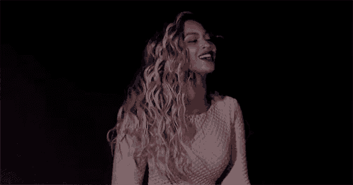 a woman with long blonde curly hair is wearing a white top and dancing in a dark room .