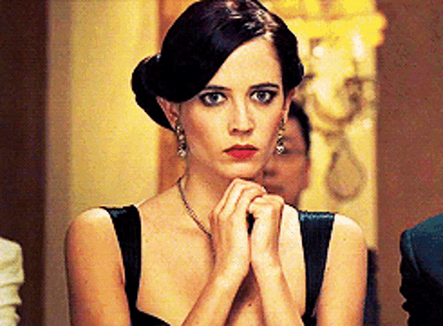 a woman in a black dress is looking at the camera with her hands folded .