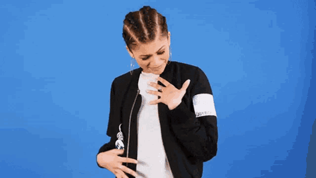 a woman in a black jacket is standing in front of a blue background and holding her hands to her chest .