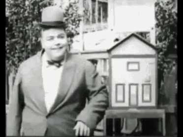 a man in a suit and top hat stands in front of a small white house