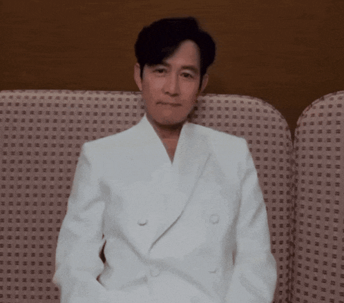 a man in a white suit sits on a couch with his hands in his pockets