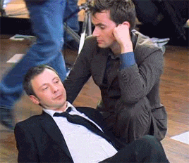 a man in a suit and tie is kneeling next to another man laying on the floor .