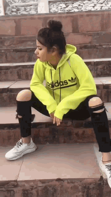 a girl wearing a neon yellow adidas hoodie and black ripped jeans is sitting on a set of stairs .