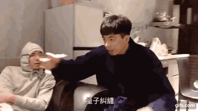 two men are sitting on a couch and one is wearing a hooded jacket .