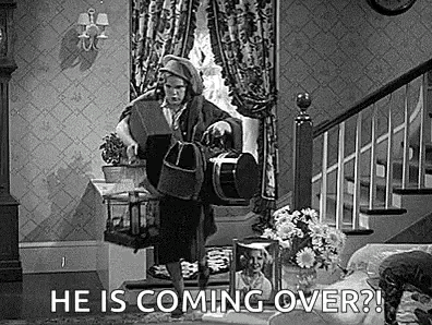 a black and white photo of a woman standing in a living room holding a bag .