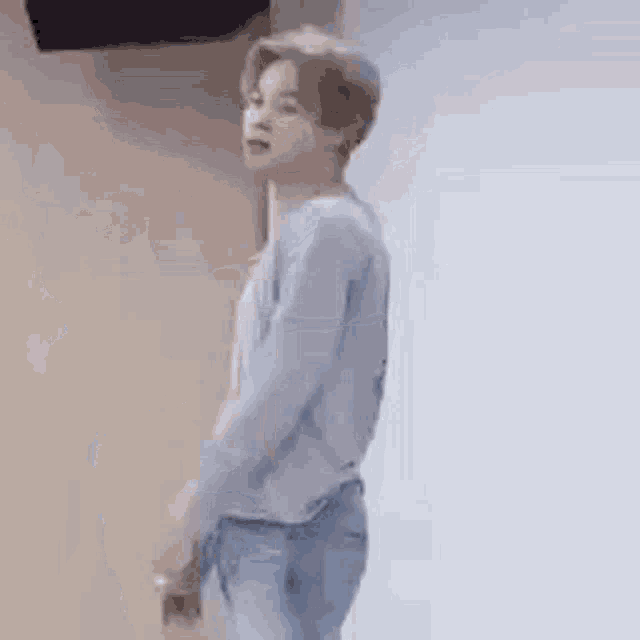 a young man in a grey sweater and blue jeans is standing in front of a white wall .