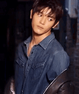a young man wearing a blue denim shirt and a black jacket is standing in a dark room .