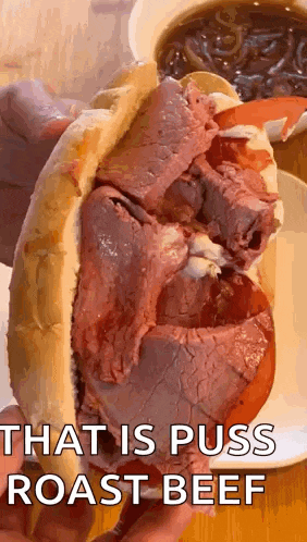 a close up of a person holding a roast beef sandwich with a cup of soup in the background .