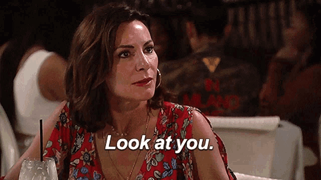 a woman in a red dress is sitting at a table with a drink and says look at you