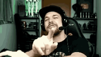 a man wearing headphones and a hat is sitting in a chair and giving the middle finger .