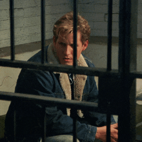 a man in a denim jacket sits in a cell behind bars