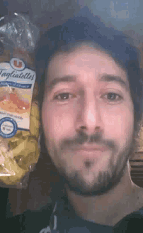 a man with a beard holds a bag of tagliatelles in front of his face