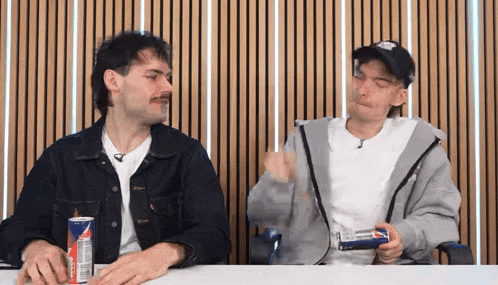 two men are sitting at a table with a red bull can in front of them