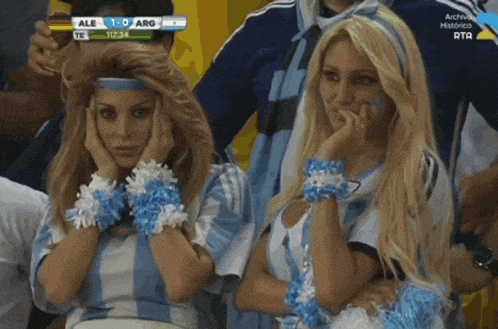 two women are watching a soccer game and the score is 1-0