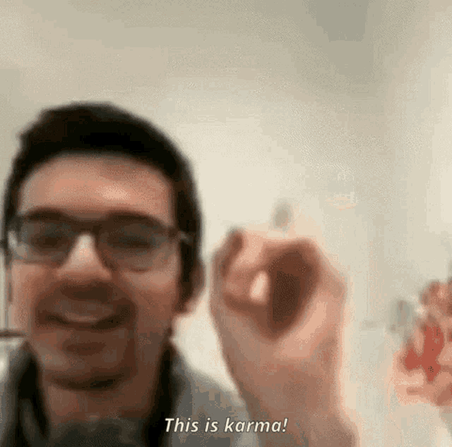 a man wearing glasses is smiling and holding something in his hand while saying `` this is karma '' .