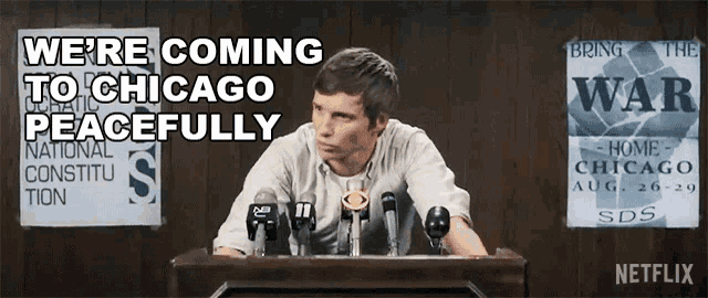 a man sitting at a podium with a sign that says " we 're coming to chicago peacefully " behind him