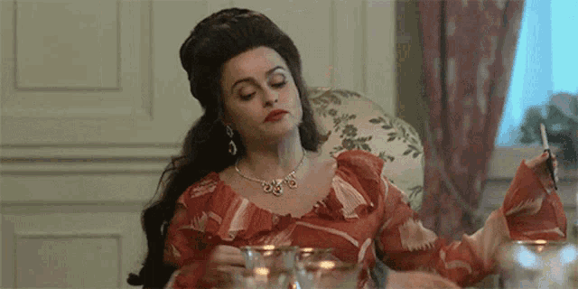 a woman in a red dress is sitting at a table with a glass of water and a cigarette in her hand .