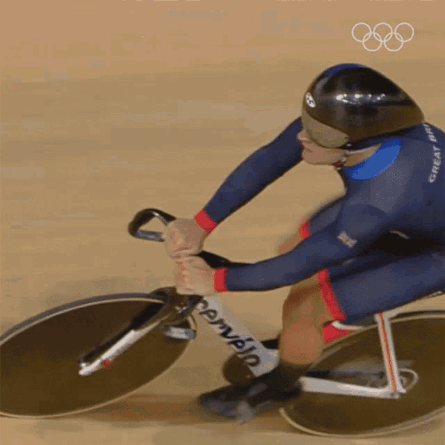 a man wearing a helmet and sunglasses is riding a bike on a track with the olympic rings in the background