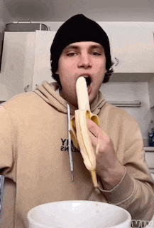 a man wearing a beanie is eating a banana from a white bowl