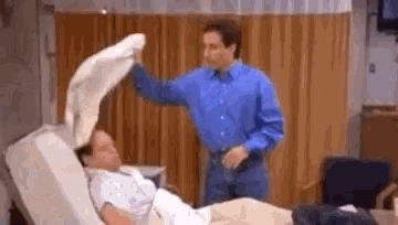 a man in a blue shirt is putting a blanket on a man in a hospital bed .