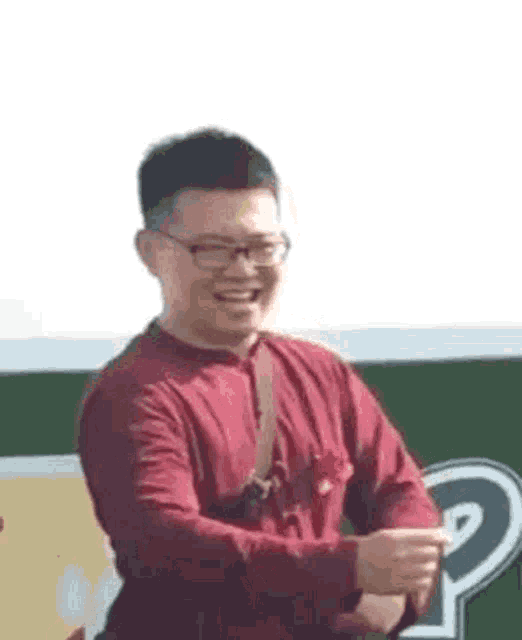a man wearing glasses and a red shirt is standing in front of a green sign and smiling .