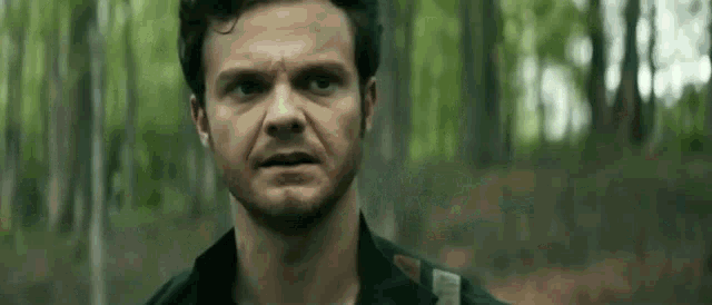 a man with a beard and a black shirt is standing in the woods looking at the camera .