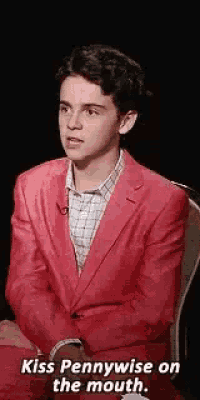 a young man in a red suit is sitting in a chair and saying `` kiss pennywise on the mouth . ''