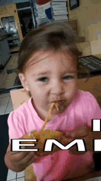 a little girl in a pink shirt is eating a hamburger with the word em on it .