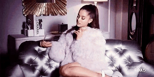 ariana grande is sitting on a couch in a living room wearing a fur coat and choker .