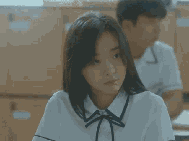 a girl in a school uniform is sitting in a classroom with a boy in the background
