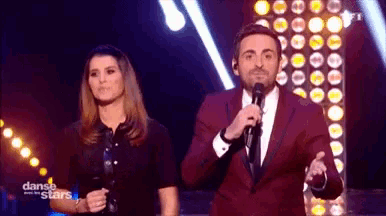 a man in a red suit stands next to a woman holding a microphone on a dance with the stars stage .