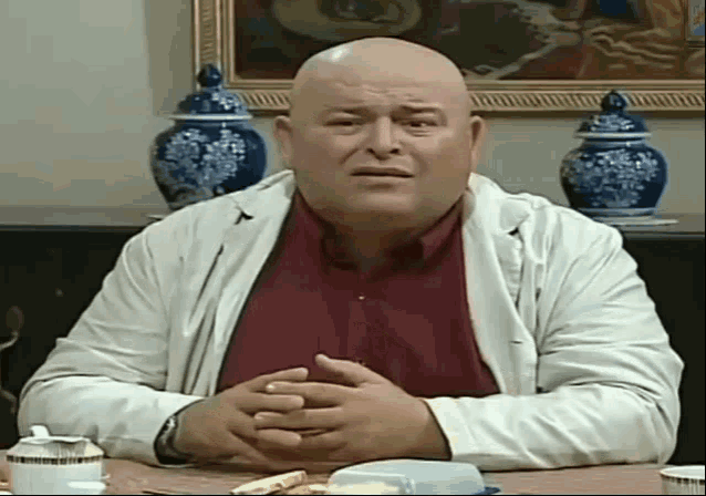a bald man is sitting at a table with his hands folded in front of a painting .