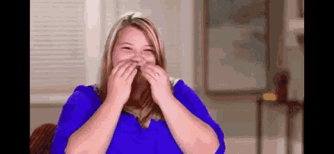 a woman in a blue shirt is covering her mouth with her hands while smiling .