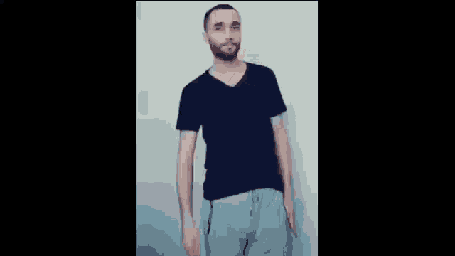 a man with a beard wearing a black t-shirt and grey sweatpants is standing in front of a wall .