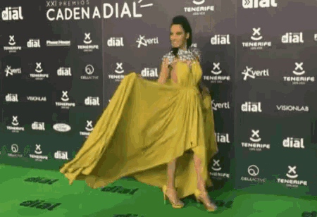a woman in a yellow dress stands on a green carpet with cadena dial written on the wall behind her
