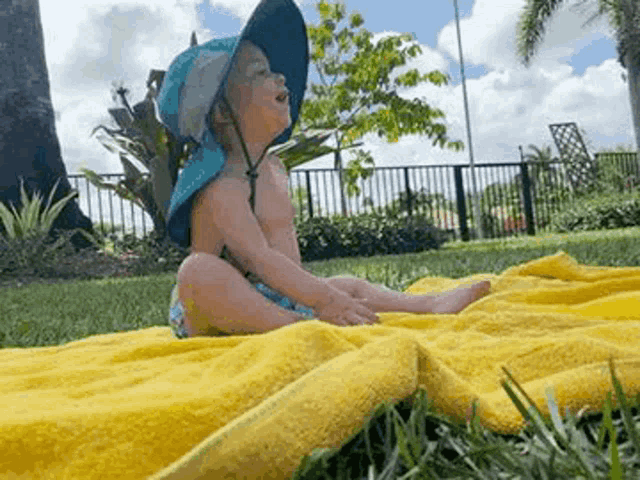 a baby wearing a blue hat is sitting on a yellow towel in the grass .
