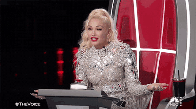 a woman in a silver dress is sitting in front of a podium with #thevoice on the bottom