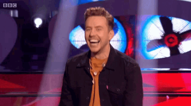 a man is laughing on a stage with a bbc logo in the background