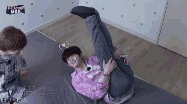 a boy in a pink shirt is laying on a bed with his legs crossed while another boy looks on .