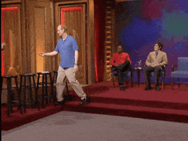 a man in a blue shirt stands on a stage while two men sit in chairs