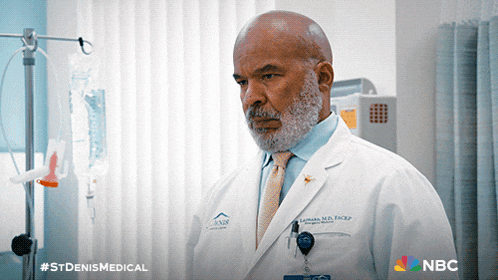 a doctor in a white coat and tie is standing in a hospital room with a nbc logo behind him