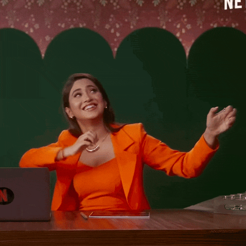 a woman in an orange suit is sitting at a desk in front of a laptop